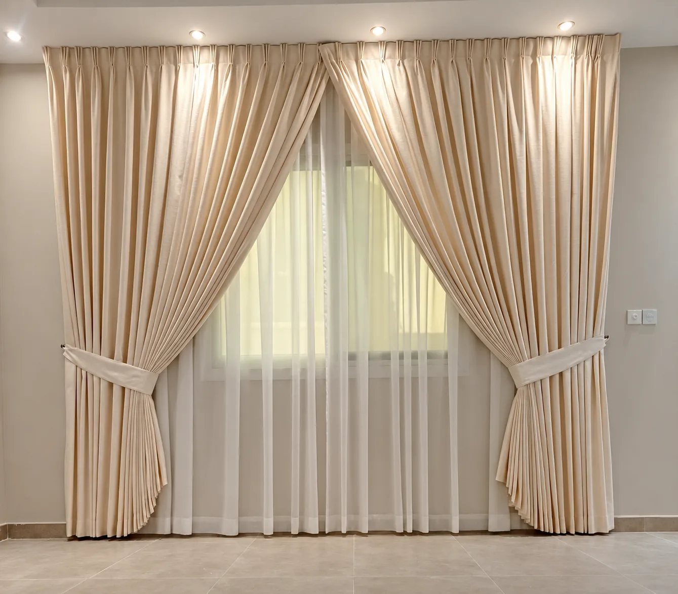 Cream triple pleat curtains with tiebacks over a sheer, fixed to the ceiling